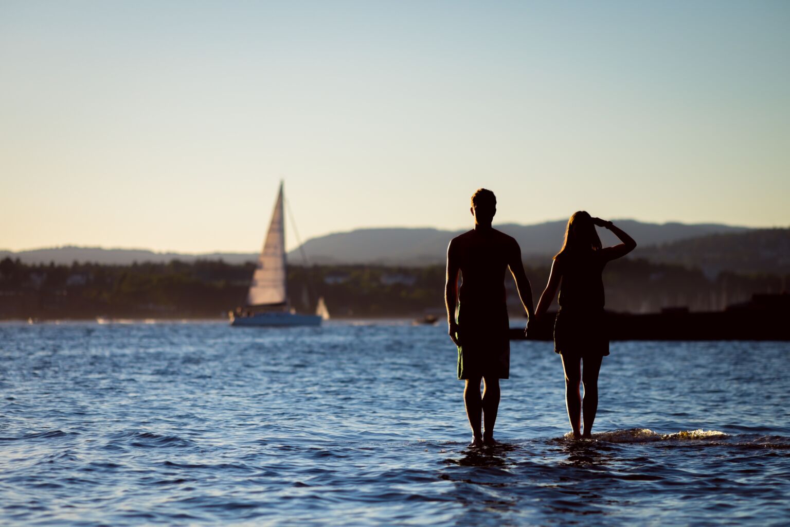 Två människor som står i vattnet och håller varandra i händerna, en segelbot syns i horisonten