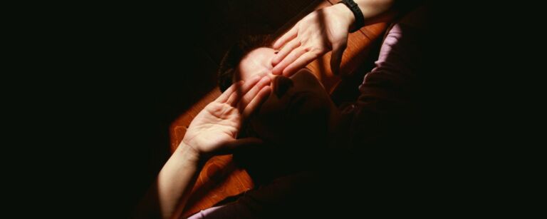Mustan keskellä avautuu valokeila, jossa ihminen makaa lautalattialla kämmenet ylöspäin asetettuna silmille. Henkilöllä on vaalea iho.